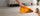 A person's hand holding a yellow-orange carpet cleaner pad over a beige carpet, lifting a stained section to show cleaning capability. A coffee cup with spilled coffee is on the right, and a sofa is in the background.